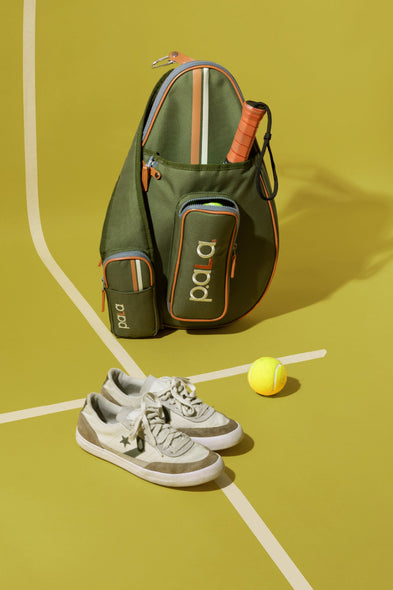 An olive green Pala pickleball bag with orange and white accents, styled on a yellow tennis court background with white court lines. A tpickleball racket is tucked into the top pocket, a pair of white and gray sneakers is in the foreground, and a yellow tennis ball sits to the right.